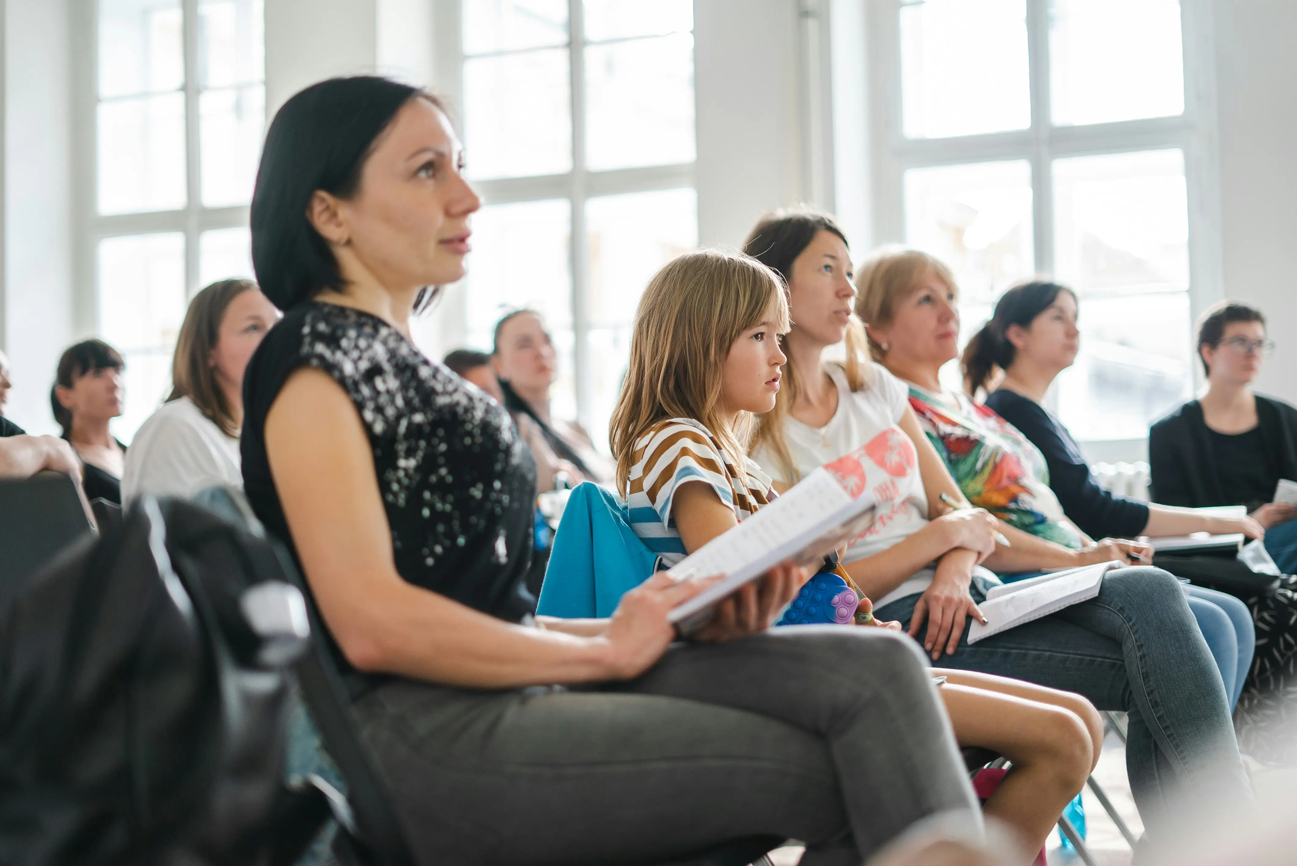 A mixed group of people sit on chairs in rows facing forwards listing or watching an event take place